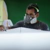 Artisan skillfully shaping a surfboard with protective gear in a workshop environment.