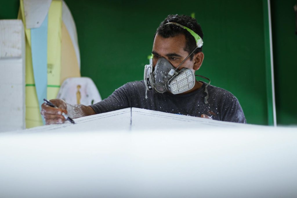 Artisan skillfully shaping a surfboard with protective gear in a workshop environment.